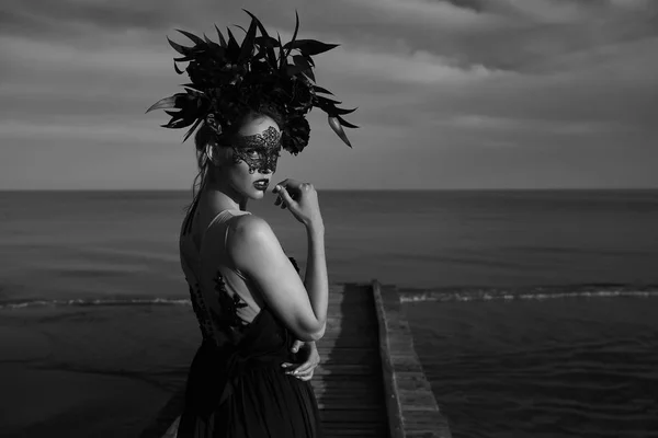 感情的な美人顔に黒レースのマスクと頭に大きな花の冠の肖像画を劇的な黒と白の木製の桟橋の上に立ちます 暗い雲と海を背景に — ストック写真