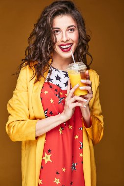 Koyu kahverengi dalgalı saçlı, güzel gülümseyen kadın neşeli parlak portre renkli yıldız yazdırma elbiseli, sarı ceket, elinde portakal suyu ile giyinmiş. Turuncu arka plan  