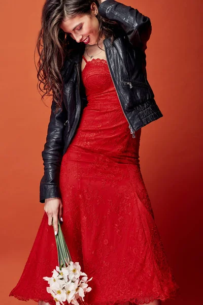 ウェーブのかかった髪と美しい笑顔ブルネットの女性服を着た赤レース ドレス 黒革のジャケットがオレンジ色の背景とスタジオで水仙の束でポーズ — ストック写真