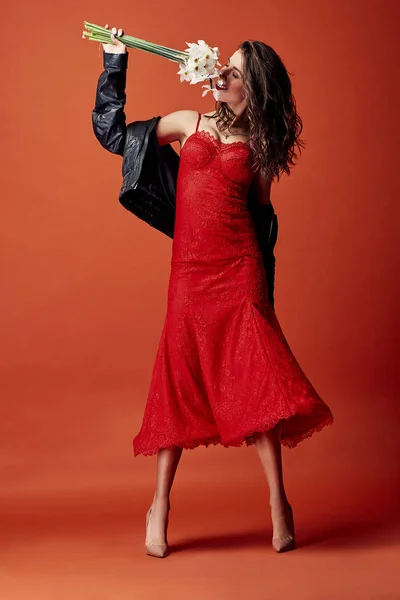 黒革ジャケットとベージュ ハイヒールのオレンジ色の背景とスタジオで水仙の束でポーズのセクシーな赤レース ドレスに身を包んだ陽気なブルネットの女性 — ストック写真