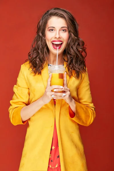 Colorful portrait of beautiful woman with dark brown hair and eyes, dressed in bright star print dress and yellow jacket, with orange juice in hands, posing in studio with dark orange background