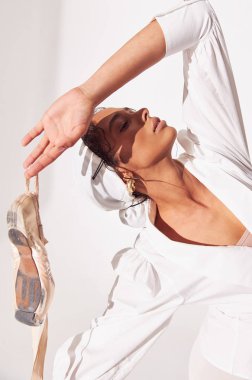 Beyaz arka planda koyu tenli balerin portresi fotoğrafı, beyaz gömlek, bej tulum, beyaz tayt ve kollarında bej rengi noktalar ile arkasına yaslandı ve bir elini kaldırdı.