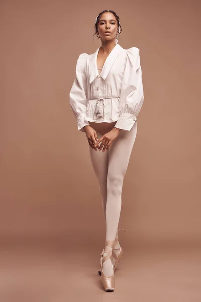full-length photo of dark-skinned ballerina on brown background wearing white shirt, beige bodysuit, white tights and beige pointes she looks into camera holding hands in front of her