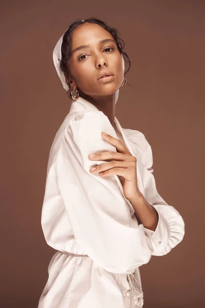 portrait of a dark-skinned girl on a brown background dressed in a white cotton shirt and a white hat she looks into the camera holds her  hands on her body she has dark eyes and dark hair