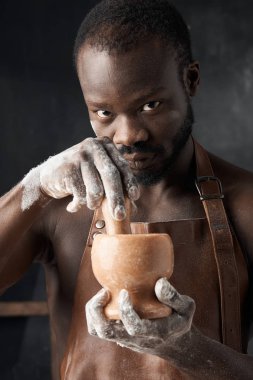 Koyu tenli, kahverengi deri önlüklü genç adam karanlık bir odada çıplak bedenin üzerinde elinde bir değirmenle kameraya bakıyor.