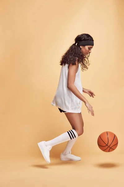 beautiful dark-skinned girl stands in full height on a yellow background in white sports shirt, black shorts, white high socks and sneakers playing with basketball ball