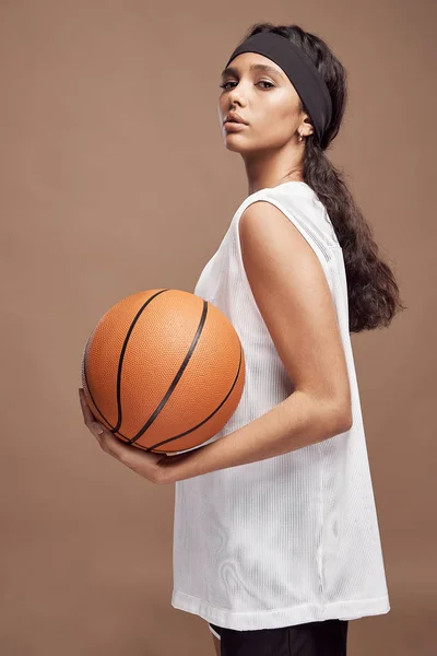 portrait of a beautiful dark-skinned girl who stands on a beige background in  white sport shirt and black shorts with a basketball ball in hand and looking into the camera