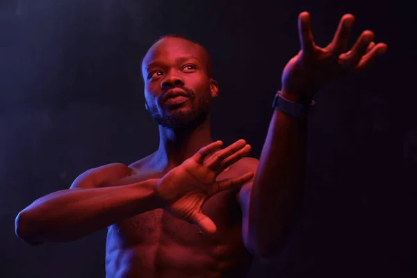 portrait of a black guy with a naked torso on a dark background with a red light raising his hands in front of him and he has a sports watch on his arm