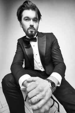 İskemlede oturan yakışıklı bir sakallı adamın siyah beyaz portresi, siyah smokin giyiyor, siyah papyonlu beyaz klasik gömlek ve kameraya bakıyor.