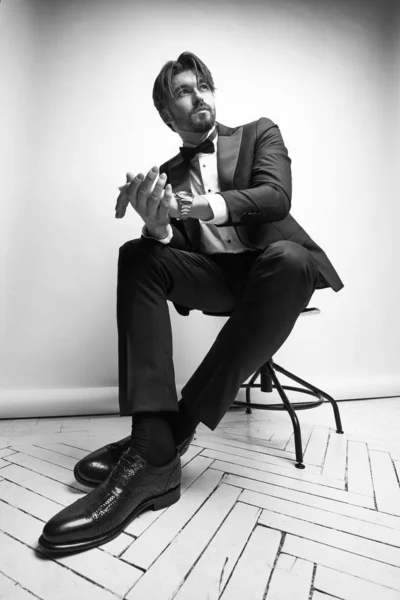 black and white portrait of a handsome young beard man, sitting on the chair, he is wearing  black tuxedo, a white classic shirt with a black bow tie and classic shoes