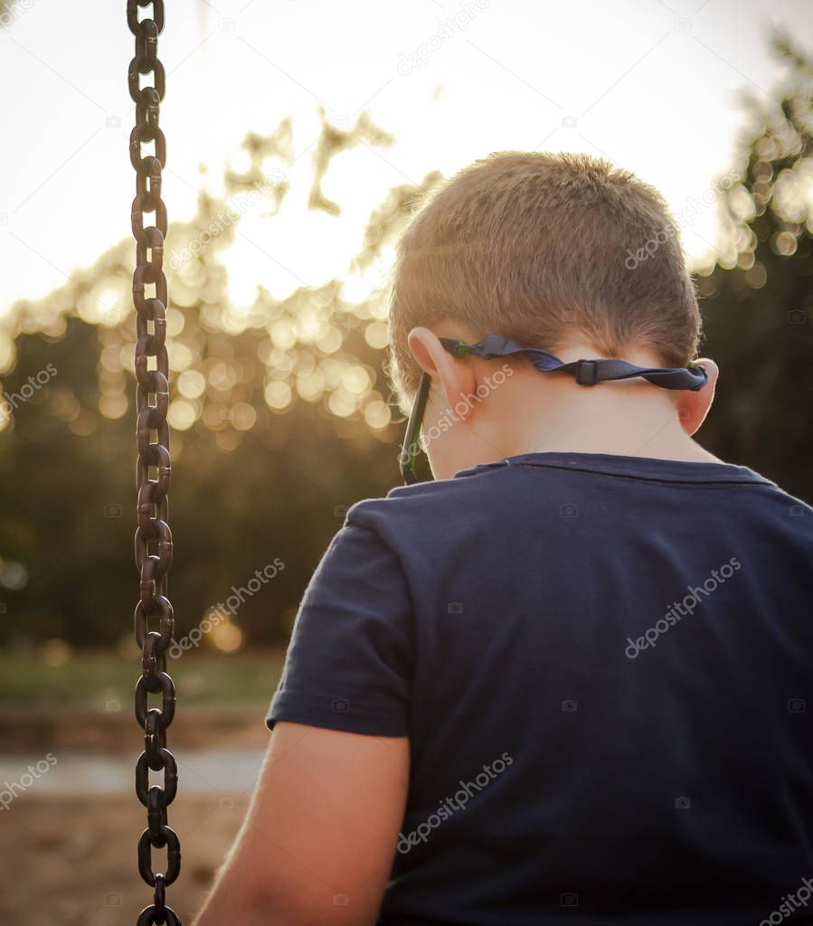Sad boy with glasses looks at the floor sitting on a swing witho