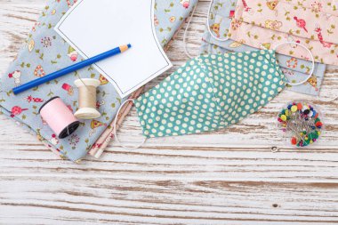 Beyaz ahşap arka planda kumaş, iplik ve aksesuarlarla testereyle kesilmiş DIY pamuk yüz maskesi, üst görünüm