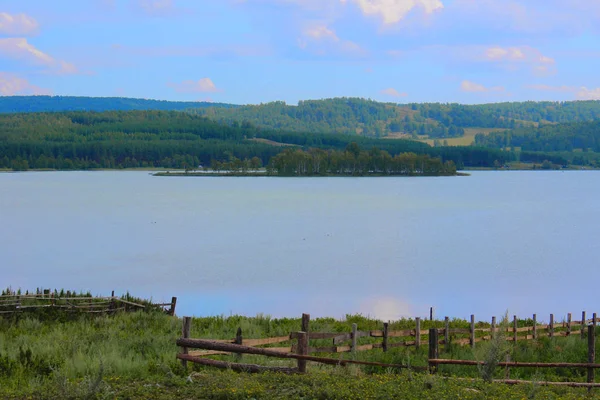 Piękny letni krajobraz błękitnego jeziora, nieba, lasu i zielonego brzegu z drewnianym płotem. Rosja, południowe Urale Baszkortostanu. — Zdjęcie stockowe