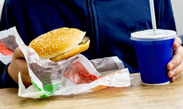 Closeup photo of hands holding a burger and a blue paper cup with a cocktail straw. A young man in a cafe snacking on junk food. Student lunch