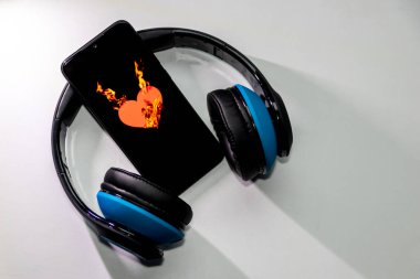 Siyah bir akıllı telefon ekranında yanan alevlerle yanan kalp sembolü ve kulaklık ve fotokopi alanı müzik ve mobil müzik ve müzik dinleme hizmetlerini gösteriyor.