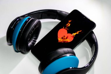 Siyah bir akıllı telefon ekranında yanan alevlerle yanan kalp sembolü ve kulaklık ve fotokopi alanı müzik ve mobil müzik ve müzik dinleme hizmetlerini gösteriyor.