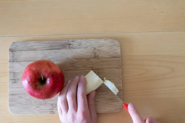 European child hands cutting fruit like red apple and banana with a sharp knife into pieces for breakfast or as healthy snack with vitamins on a wooden plank on the kitchen table as diet meal