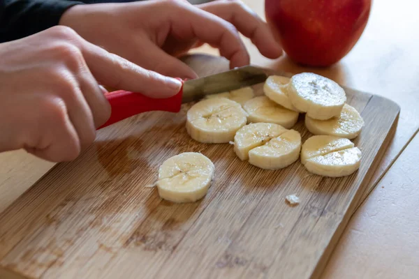 European child hands cutting fruit like red apple and banana with a sharp knife into pieces for breakfast or as healthy snack with vitamins on a wooden plank on the kitchen table as diet meal