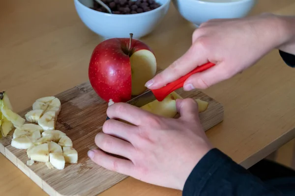 European child hands cutting fruit like red apple and banana with a sharp knife into pieces for breakfast or as healthy snack with vitamins on a wooden plank on the kitchen table as diet meal