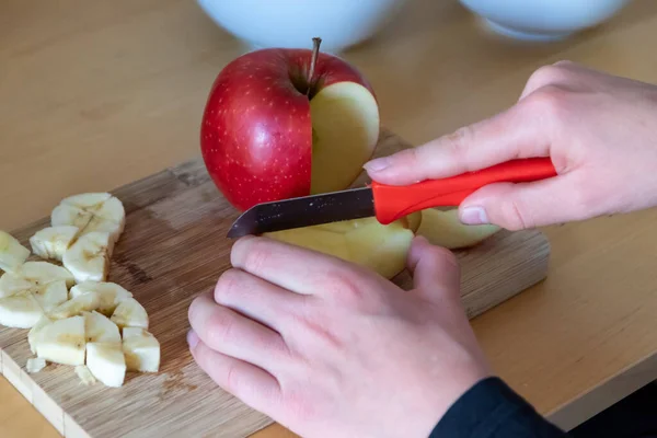 European child hands cutting fruit like red apple and banana with a sharp knife into pieces for breakfast or as healthy snack with vitamins on a wooden plank on the kitchen table as diet meal