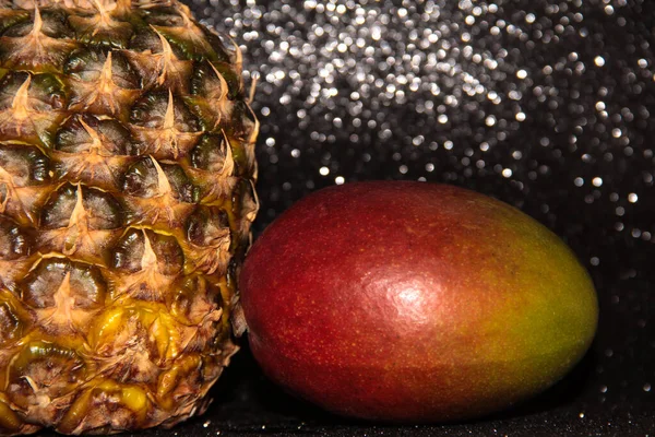 Pineapple with mango on a black shiny background. Half a pineapple and a whole mango in the skin.