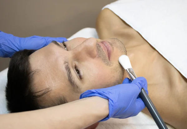 Νεαρός ξαπλωμένος σε έναν καναπέ θεραπείας προσώπου καλυμμένος με λευκή πετσέτα με ανοιχτά μάτια. χέρια φυσιοθεραπευτή κρατώντας το πινέλο προσώπου πάνω από το πρόσωπο του ανθρώπου σε ένα μπλε αποστειρωμένα γάντια — Φωτογραφία Αρχείου