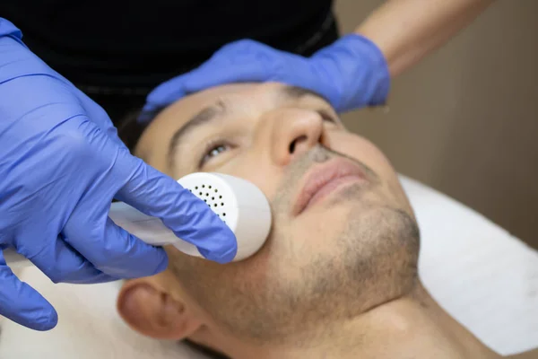 Jongeman ligt op een gezichtstherapie bank bedekt met de witte handdoek met geopende ogen. Gezichtstherapeut handen houden de gezichtsmachine boven de mens gezicht in een blauwe steriele handschoenen — Stockfoto