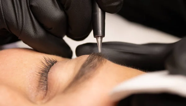 Jonge aantrekkelijke man oog close-up met de master hand in zwarte handschoenen en permanente make-up machine met een naald. — Stockfoto