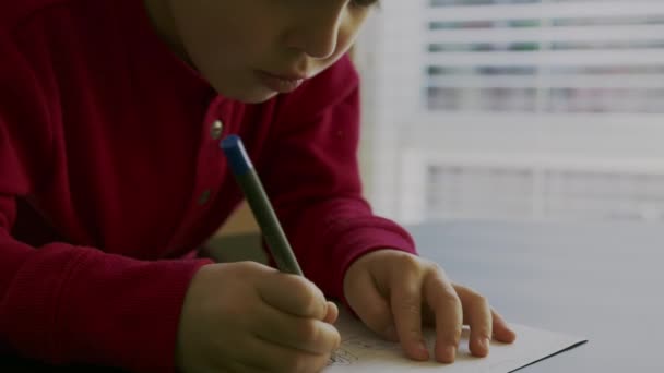 Menino em um suéter vermelho sentado em uma mesa que ele está segurando uma caneta em suas mãos e escrever ou desenhar em um pedaço de papel branco em branco na frente de uma janela brilhante no fundo . — Vídeo de Stock