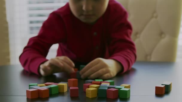 Niño pequeño en un suéter rojo está jugando con los bloques de madera de colores en una mesa delante de una ventana. . — Vídeos de Stock