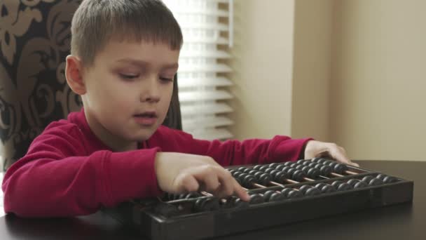 Маленький школьник в красном свитере сидит за столом и учится математике на старом счёте. счастливый ребенок учит математике на абакусе — стоковое видео