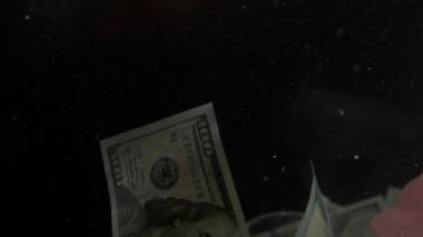 Amerikan dolarlarıyla karışan çeşitli plastik ve yiyecek çöpleri yüksek kaliteli, çevresel sorunlar ve okyanus kirliliği içeren siyah bir arka plan yavaş çekim videosunda suda yüzüyor. Yüksek kalite