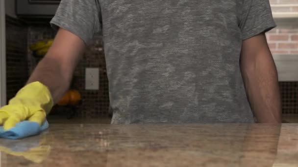 Young man in a gray T-shirt cleaning kitchen granite countertop in his house. He is wearing a bright yellow rubber glove and holds a blue rag which he is using for wiping the kitchen table with a — Stock Video