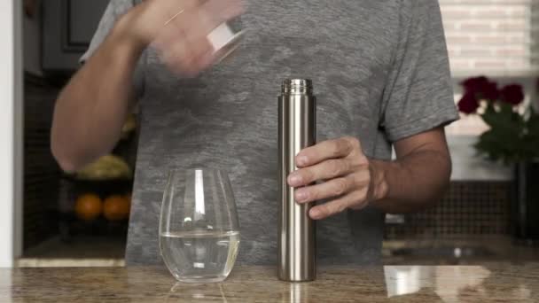 Jeune homme dans un T-shirt gris verse de l'eau dans un verre à partir d'un thermos ou une bouteille en plastique dans une belle cuisine à la mode. mode de vie sain. Un bel homme a soif. . — Video