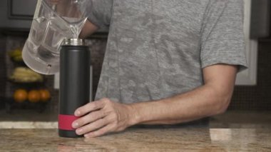 Gri tişörtlü genç adam şık bir mutfakta plastik şişeden termosa su döker. Sağlıklı bir yaşam tarzı. Yakışıklı adam susamış..