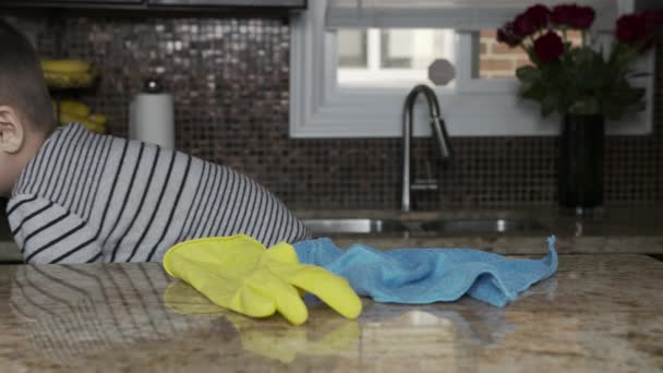 A little cute boy in a gray striped sweater sits on a high chair and helps to tidy up the kitchen, he holds a blue rag in his hand with a yellow rubber glove and wipes the kitchen table. happy — Stock Video