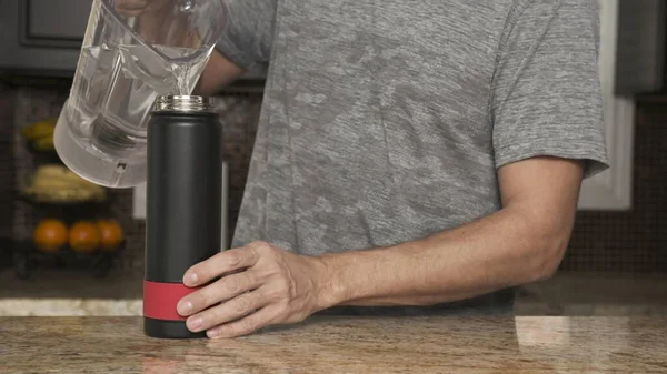 Gri tişörtlü genç adam paslanmaz çelikten bir termostan bardağa su döker ya da güzel bir mutfaktaki plastik şişeye. Sağlıklı bir yaşam tarzı. Yakışıklı adam susamış..