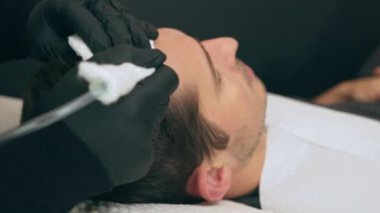 Yüksek kalite yakın çekim 4K yavaş çekim videosu. Estetisyen saç tedavisiyle koltukta yatan bir adam. Erkek tricopigmentasyon servisi. Kafa derisi mikro pigmentasyon tedavisi. Trikopigmentasyon prosedürü.
