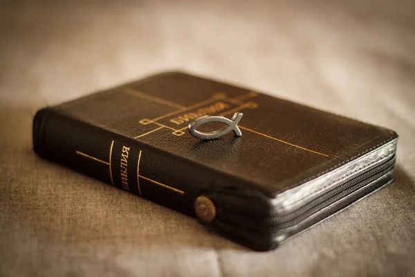 Foto de um livro Bíblia close-up em couro preto encadernação com um zíper com um peixe símbolo pingente cristão em um fundo cinza — Fotografia de Stock