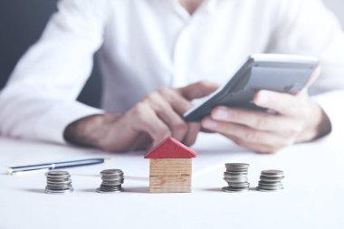 Businessman Calculating Tax In Front Of Stacked Coins And House 