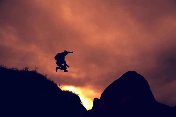 man silhouette jump happy from cliff over gap sunset