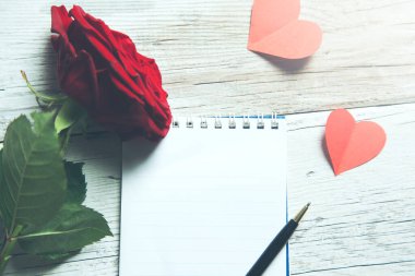 Not defterli kırmızı gül ve tahta masa arkasında kalpler.