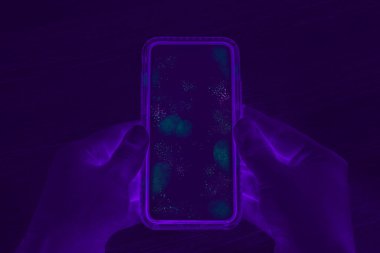 Kirli dokunmatik ekranlı cep telefonu tutan eller - UV Blacklight bulaşıcı bakterileri ve mobil akıllı telefon ekranında zararlı mikropları açığa çıkarıyor - hastalık, korona virüsü ve hijyen konsepti