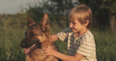 Doğada köpeği olan bir çocuk. 7 yaşında köpek besleyen bir çocuk Alman çoban köpeği besliyor. Sahada köpekle gülümseyen mutlu çocuk. Küçük çocuk çayırda evcil hayvanıyla oynuyor. Yavaş çekim. 
