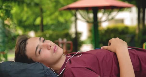 Портрет Подростка Наушниках Лежащего Скамейке Парке Расслабляющего Подросток Слушает Музыку — стоковое видео