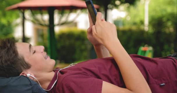 Portrait Happy Teenage Boy Mobile Phone Lying Bench Park Smiling — Stockvideo