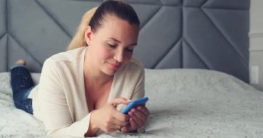 Evde cep telefonu kullanan yetişkin bir kadın. Yetişkin, güzel bir kadın akıllı telefondan mesaj yazıyor. Mutlu gülümseyen kadın yatağında uzanmış cep telefonu kullanıyor. Neşeli kadın akıllı telefonu tutuyor..        