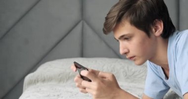Ergen, akıllı telefonla, evde oyunlar oynuyor. Yakışıklı çocuk koltukta uzanarak cep telefonu kullanıyor. Oyuncu cep telefonuyla oynar. Yaşam tarzı konsepti. Gerçek zamanlı görüntüler