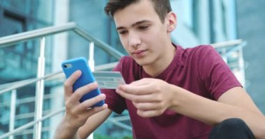 Gençler internet üzerinden kredi kartı ve cep telefonu ile alışveriş yapıyorlar. Gülümseyen genç adam açık havada, banka kartlı cep telefonuyla online alışveriş yapıyor. Çocuk akıllı telefon ve kredi kartı kullanıyor. Gerçek zamanlı..
