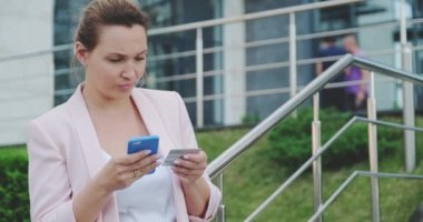 Yetişkin bir kadın kredi kartı ve cep telefonu kullanarak online alışveriş yapıyor. Gülümseyen kadın açık havada, banka kartlı cep telefonuyla online alışveriş yapıyor. Bayan akıllı telefon ve kredi kartı kullanıyor. Gerçek zamanlı.